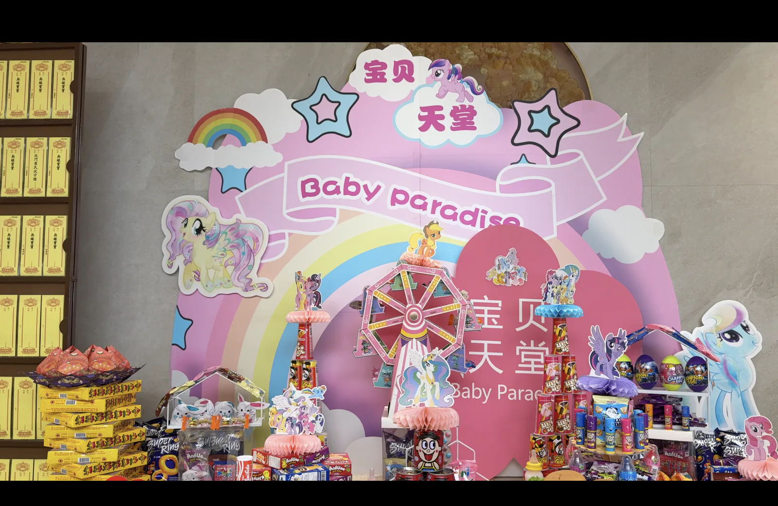 The pink Baby Paradise 宝贝天堂 backdrop at the Seventh Month prayer services at Nirvana, with a rainbow, cartoon ponies and a paper ferris wheel above tables of candies and snacks