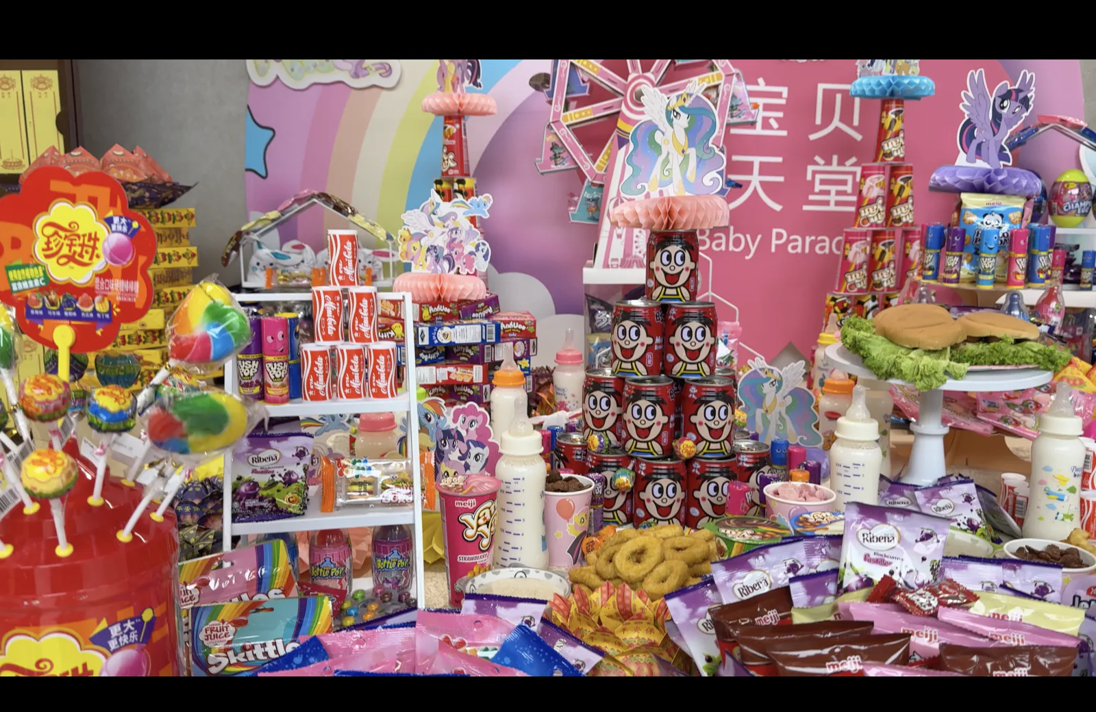 Offerings at Baby Paradise: feeding bottles of milk, lollipops, chocolate biscuits, sweets and stacked tins of a childhood soft drink before the pink backdrop